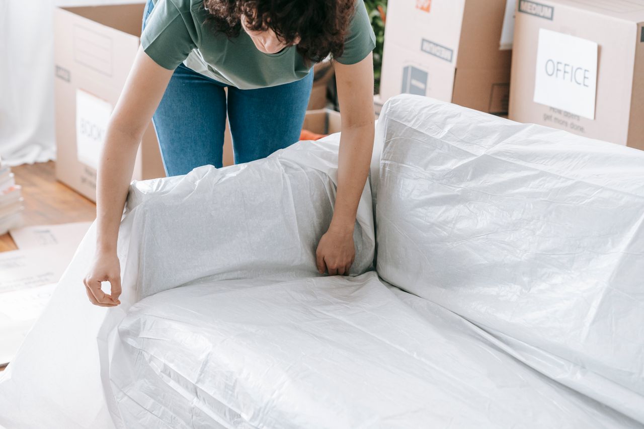 Une femme enveloppe soigneusement un canapé dans un matériau de protection tout en préparant des cartons dans une pièce lumineuse.